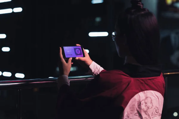 Вид сзади девушки, держащей смартфон с веб-магазином на улице с неоновым светом вечером, город будущего — стоковое фото