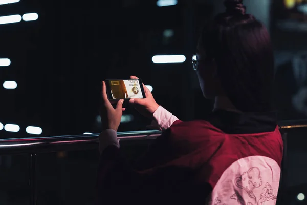 Vista posteriore di ragazza che tiene smartphone con i biglietti apparecchio on-line in strada con luce al neon in serata, città del concetto futuro — Foto stock