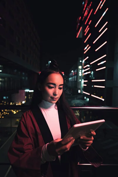 Привлекательная азиатская девушка в кимоно с помощью планшета на улице с неоновым светом вечером, город будущей концепции — стоковое фото