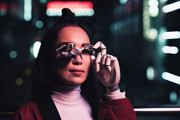 Привлекательная азиатская девушка с футуристическим протезом рук и глаз на улице вечером, город будущей концепции — стоковое фото