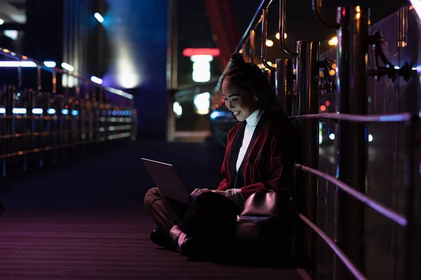 Souriant attrayant asiatique fille dans kimono assis et en utilisant ordinateur portable sur la rue avec néon lumière, ville de concept futur — Photo de stock