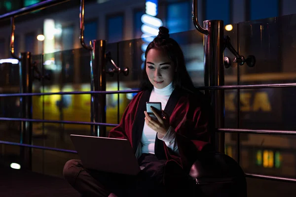 Hermosa chica asiática en kimono sentado y utilizando el ordenador portátil con teléfono inteligente en la calle con luz de neón, ciudad del concepto futuro - foto de stock