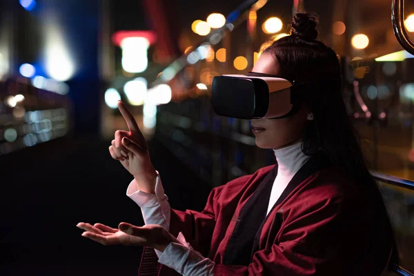 Attraente asiatico ragazza utilizzando virtuale realtà auricolare e puntando su qualcosa su strada in sera, città del futuro concetto — Foto stock