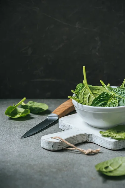 Organic fresh spinach leaves in white bowl on cutting board near metal knife — Stock Photo
