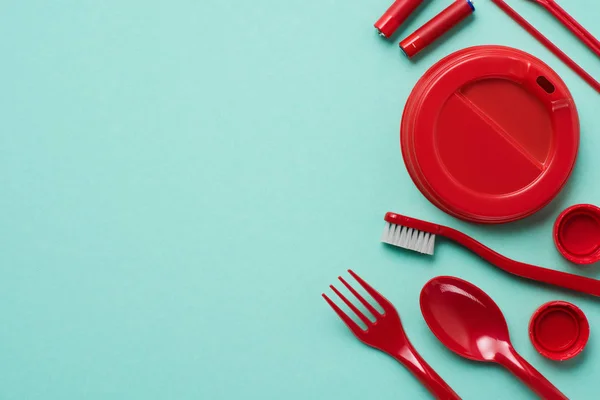 Top view of red plastic bottle caps, fork, spoon, lid for drink, batteries and toothbrush on blue background — Stock Photo