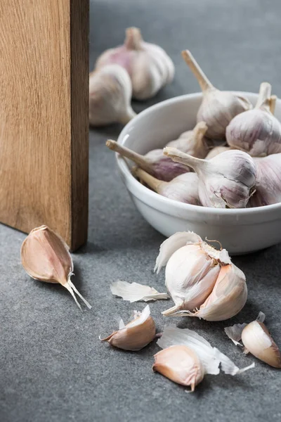 Ajo en tazón de cerámica, varios dientes pelados y tabla de cortar de madera sobre fondo gris - foto de stock