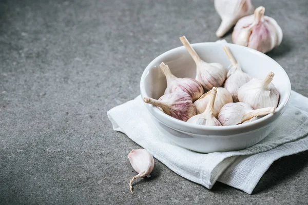 Cabezas de ajo maduras en tazón de cerámica sobre servilleta blanca y clavo pelado sobre fondo gris - foto de stock