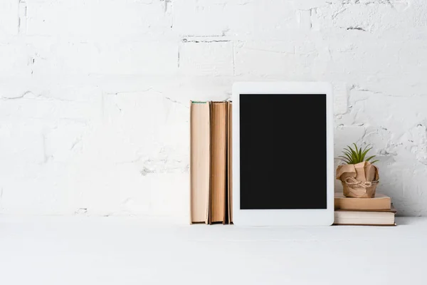 Tablet digitale con schermo bianco, pianta in vaso e libri vicino alla parete di mattoni bianchi — Foto stock