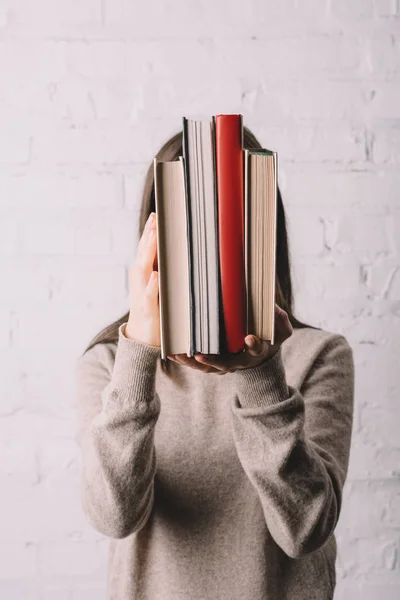 Jovem mulher escondendo rosto atrás de livros perto de parede de tijolo branco — Fotografia de Stock