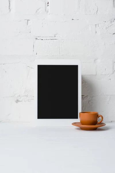 Vista de primer plano de la tableta digital con pantalla en blanco y taza de café cerca de la pared de ladrillo blanco - foto de stock