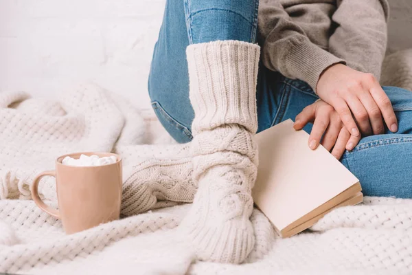 Plan recadré de fille en chaussettes tricotées assis avec livre sur couverture chaude — Photo de stock