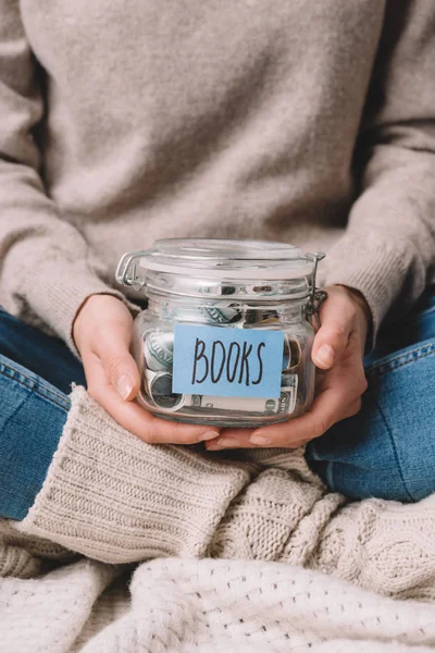 Tiro recortado de menina segurando frasco de vidro com poupança e livros de etiquetas — Fotografia de Stock