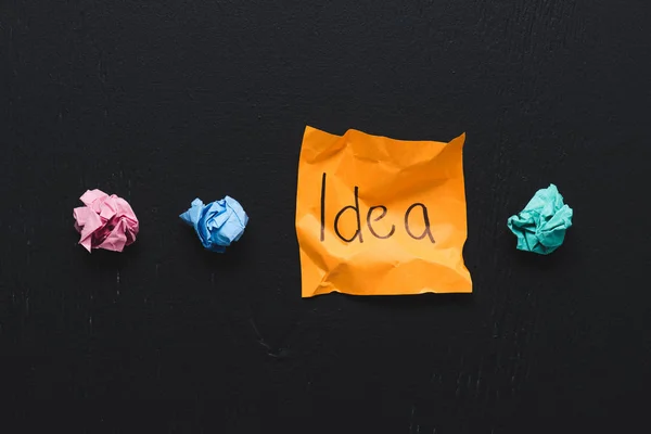 Vista superior de la palabra 'idea' escrita en nota adhesiva con bolas de papel arrugadas de colores sobre fondo negro, concepto de ideas - foto de stock