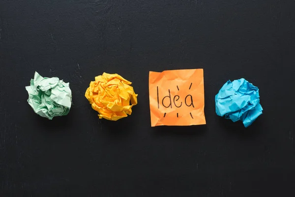 Vista dall'alto della parola 'idea' scritta su nota appiccicosa con palline di carta spiegazzate colorate su sfondo nero, concetto di idee — Foto stock
