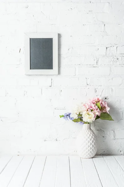 Blank frame on white brick wall with bouquet in ceramic vase on rustic wooden table — Stock Photo