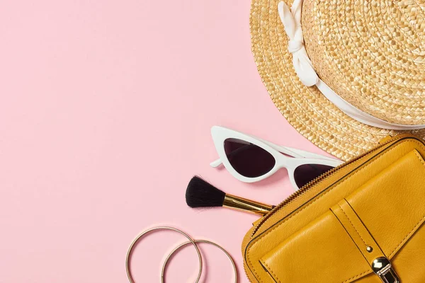 Vista superior de sombrero de paja, pulseras, cepillo cosmético, gafas de sol y bolso amarillo sobre fondo rosa - foto de stock