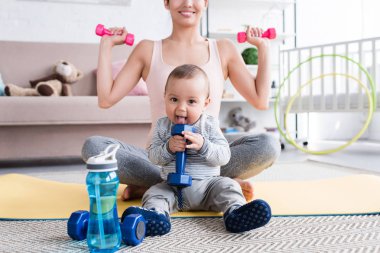 Kırpılan atış dumbbells ile yoga üzerinde otururken yapıyor gülümseyen uygun anne evde küçük çocuğuyla birlikte mat