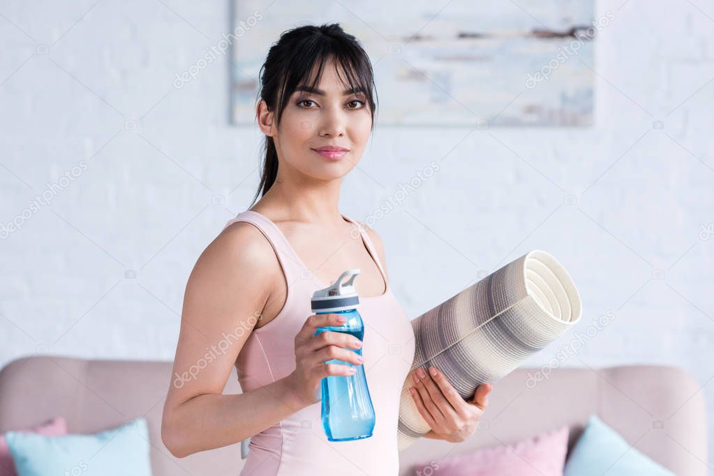 smiling young woman with rolled yoga mat and bottle of water looking at camera at home