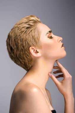 side view of attractive woman with golden glitter on face and short hair touching neck isolated on grey