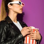 Красива молода жінка в 3d окулярах тримає коробку попкорну і дивиться ізольовано на фіолетовий