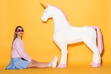 asian female model in sunglasses sitting on floor and decorative unicorn on yellow background  clipart