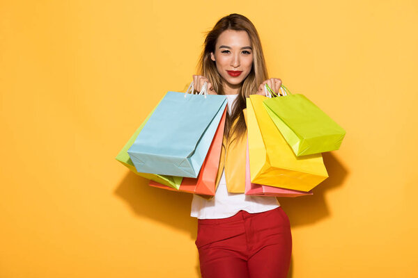 female asian shopper with paper bags standing on yellow background 