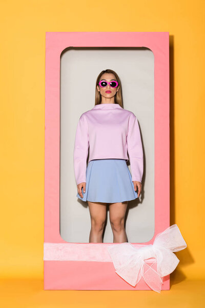 young stylish female model in sunglasses in decorative box with bow on yellow background 