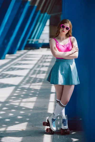 beautiful girl in sunglasses, denim skirt and roller skates standing with crossed arms and looking at camera