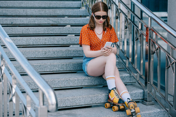 beautiful girl in sunglasses and roller skates sitting on stairs and listening music with smartphone  