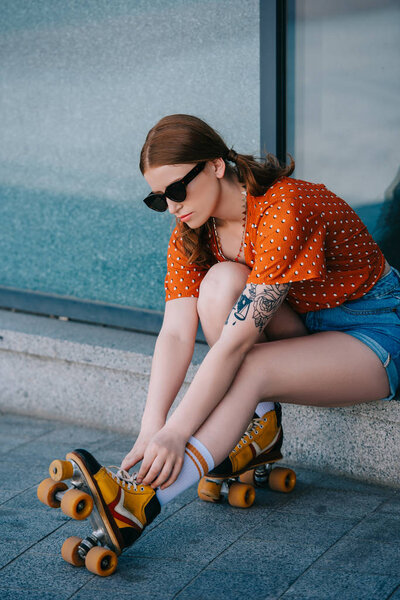 stylish girl in sunglasses wearing roller skates while sitting on street