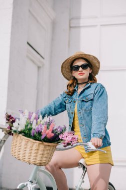 attractive girl in hat, sunglasses and denim jacket looking at camera and riding bicycle on street  clipart