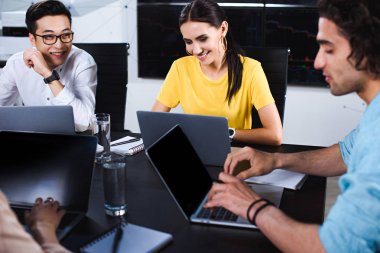 iş ortağı masada modern ofis dizüstü bilgisayarlar ile konuşurken çok kültürlü grup gülümseyen 