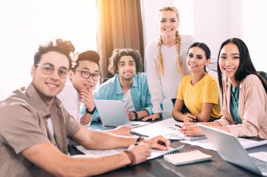 Bir grup güler yüzlü çok kültürlü iş ortağı modern ofiste toplantı sırasında dizüstü bilgisayarlarla ve kağıtlarla masada oturuyor. 