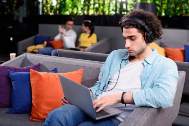 young businessman in headphones sitting on couch with laptop at modern coworking office  clipart