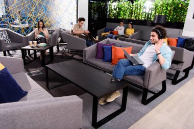 dizüstü bilgisayar ile kanepede oturan ve el sallayarak modern coworking ofiste Kulaklığımda genç işadamı 