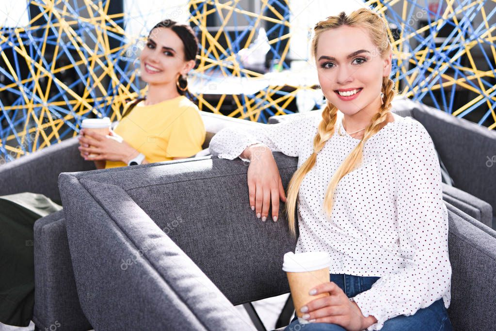 smiling businesswomen with paper cups of coffee at modern coworking office 