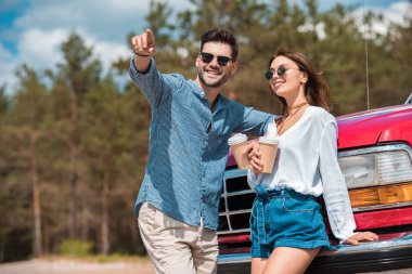couple of travelers with disposable cups of coffee standing at car, man pointing somewhere clipart