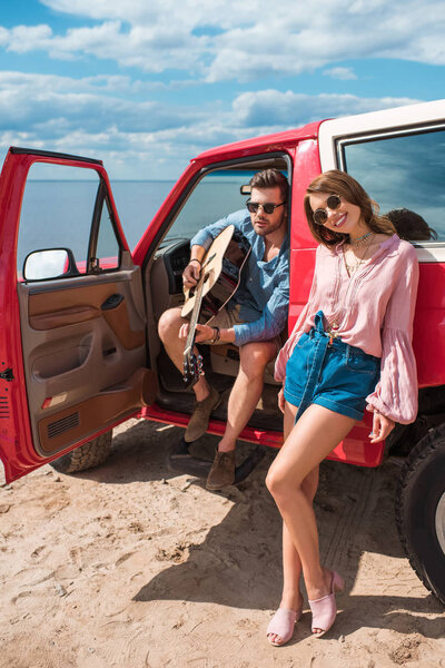 young couple with acoustic guitar near car on road trip