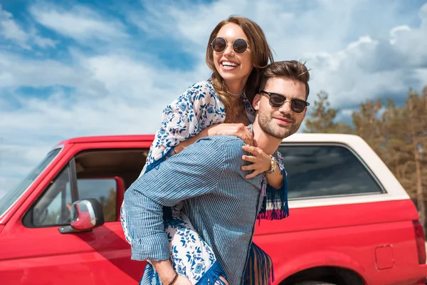 Joven Sonriente Pareja Gafas Sol Piggybacking Cerca Rojo Coche — Foto de Stock