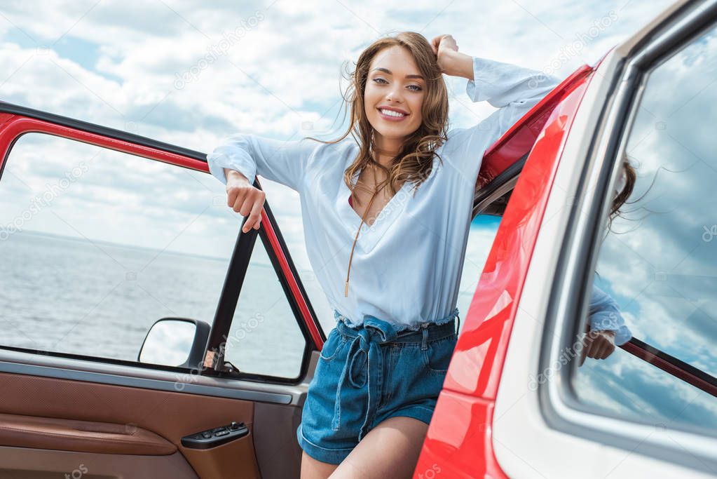 cheerful beautiful woman standing at car during road trip near the sea