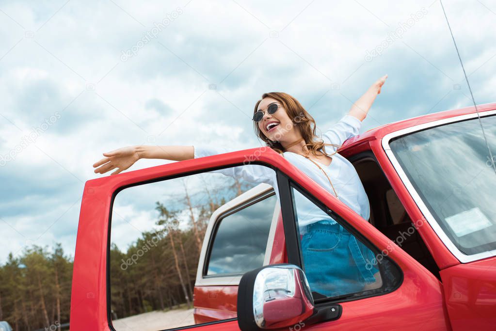 excited young woman in sunglasses posing near red car during trip