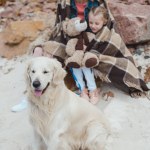 Mamma och dotter i filt sitter med hund på stranden