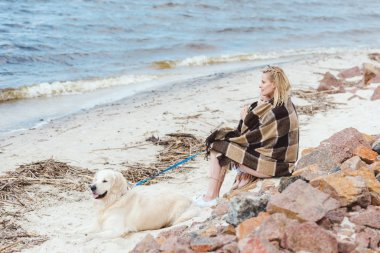 sarışın kadın ile köpek deniz kıyısında oturan battaniyeye sarılmış 