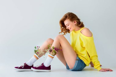 side view of beautiful smiling girl with flowers in socks sitting isolated on grey clipart