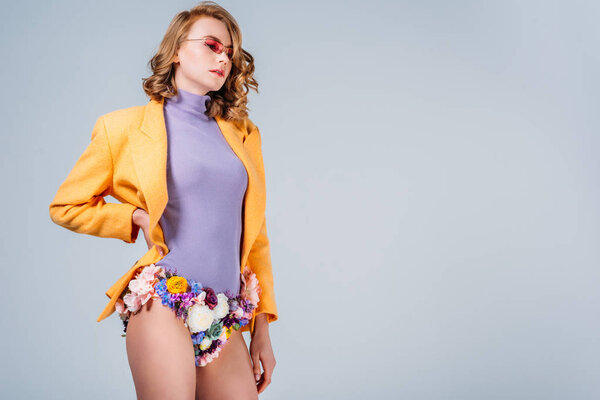 stylish girl in sunglasses and panties made of flowers standing with hand on waist isolated on grey