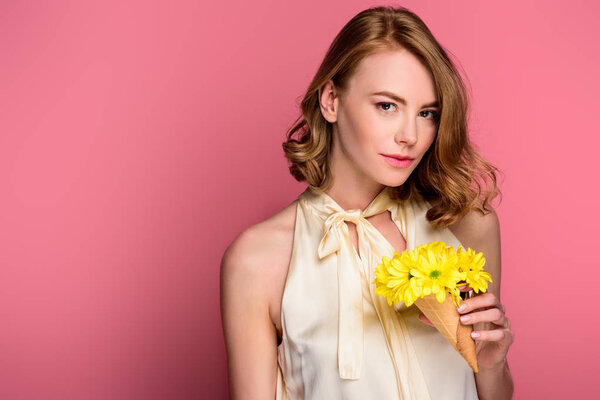 girl holding ice cream cone with yellow flowers and looking at camera isolated on pink