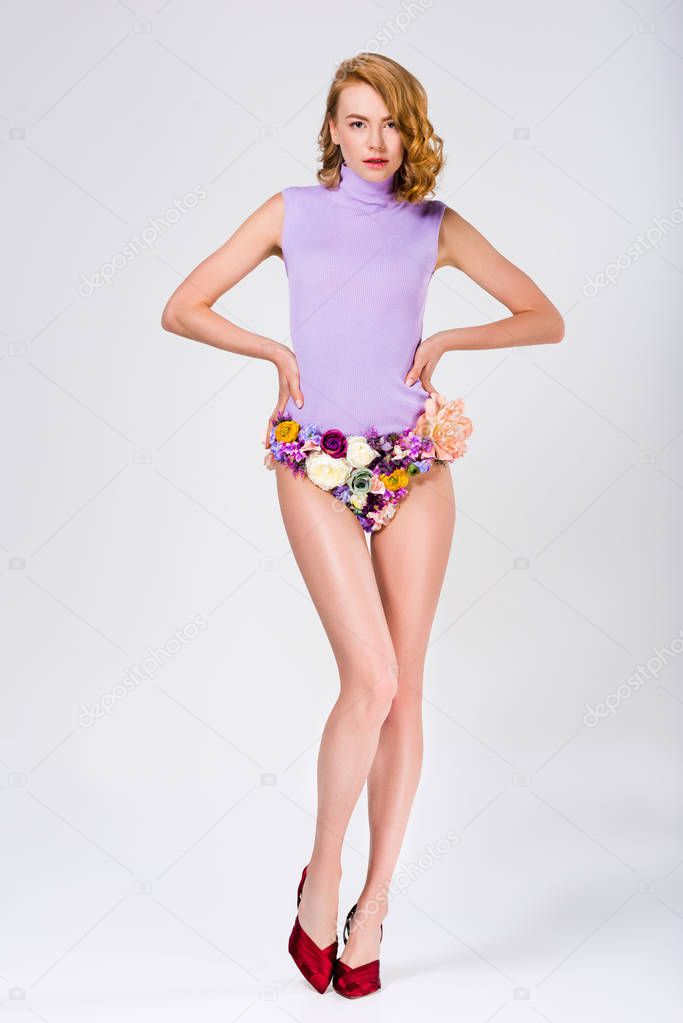 full length view of beautiful girl in panties made of flowers and high heeled shoes standing with hands on waist and looking at camera on grey