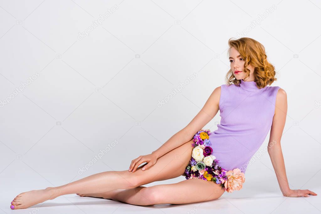 beautiful barefoot girl in panties made of flowers sitting and looking away on grey