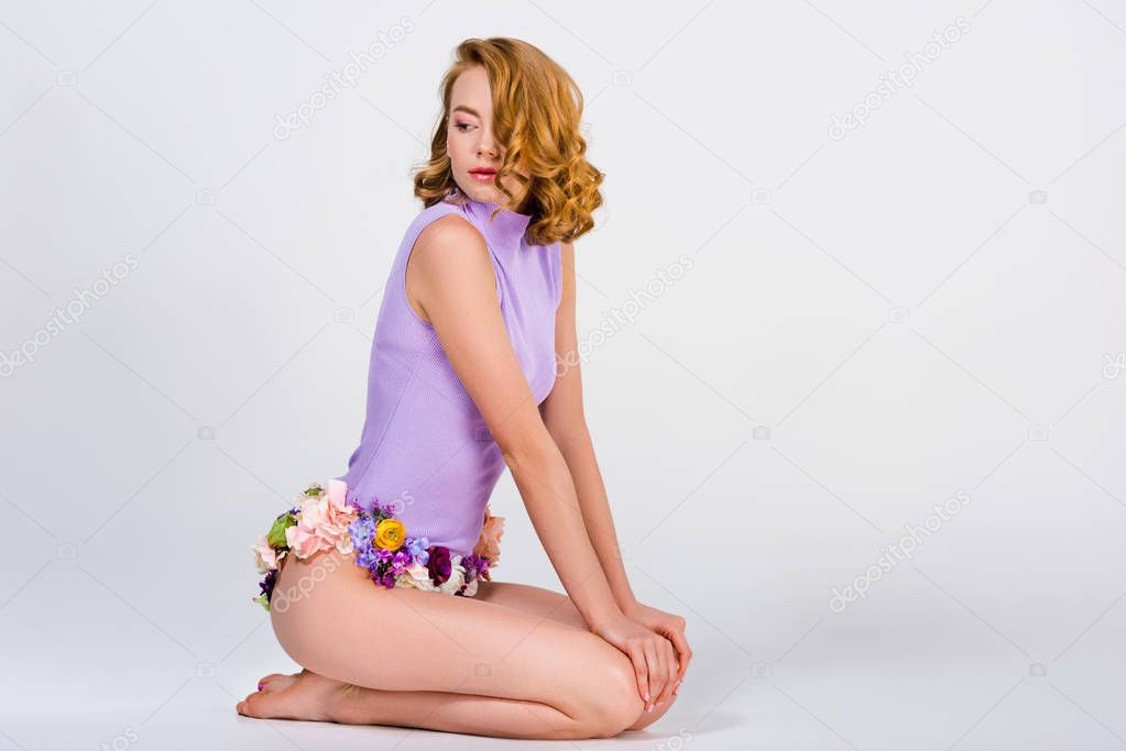 beautiful barefoot girl in panties made of flowers sitting and looking away isolated on grey