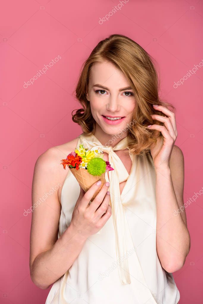 beautiful young woman holding wafer cone with flowers and smiling at camera isolated on pink 